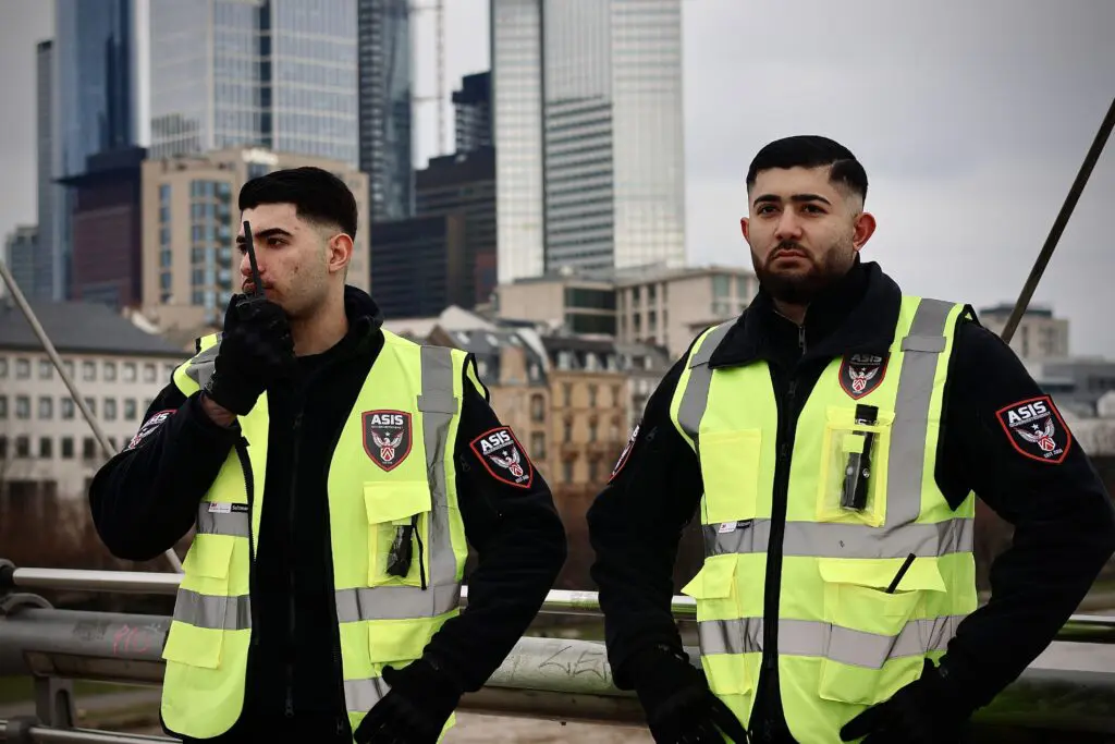Zwei ASIS Sicherheitsdienstmitarbeiter führen eine Patrouille auf einer Messe durch, im Hintergrund die Skyline der Stadt