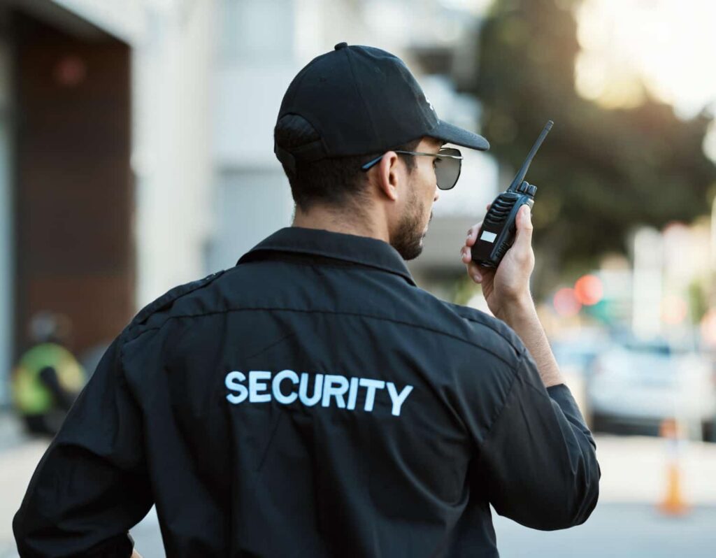 Sicherheitsdienst Hof – Ein Sicherheitsbeamter mit Funkgerät und Kappe überwacht konzentriert ein urbanes Gebiet bei Tageslicht.